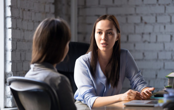 Female professional wearing a button up shirt talking to another woman in an office.