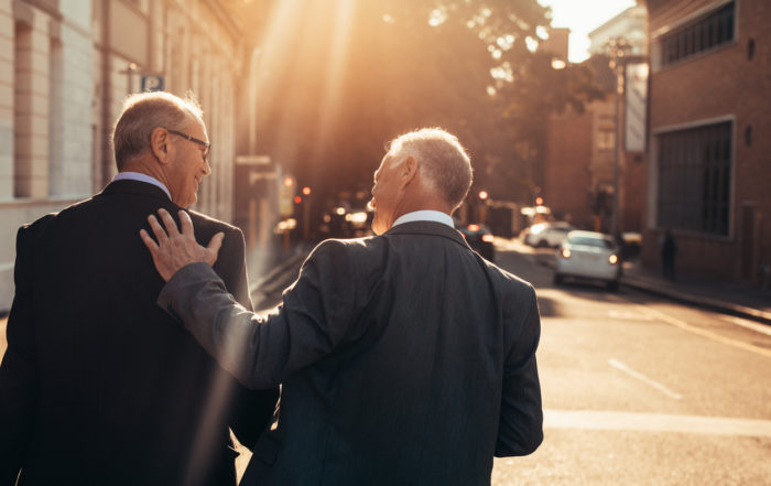 Two older men walking down the street at sunset smiling at each other.